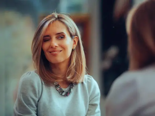 Frau mit kinnlangen Haaren zwinkert und lächelt. Sie trägt einen grauen Pullover und eine auffällige Halskette.