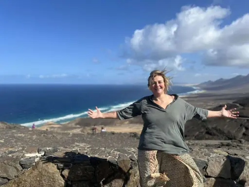 Donna sorridente con le braccia aperte su un paesaggio costiero, un'espressione di gioia che contrasta la solitudine interiore.