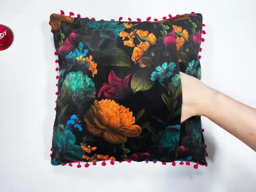Ett sammetslent kuddfodral med en mörk blommig design och en lekfull kant av små röda bollar. En hand sticker fram ur en ficka på kudden.