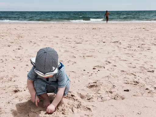 Kleines Kind mit Hut spielt im Sand am Strand der Algarve. Im Hintergrund läuft ein Mann ins Meer. Ein perfekter Tag für Algarve mit Kindern.