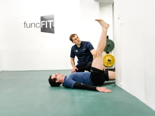 Ein Trainer hilft einem Mann beim Oberschenkel dehnen. Der Mann liegt auf dem Rücken, das Bein gestreckt an der Wand.
