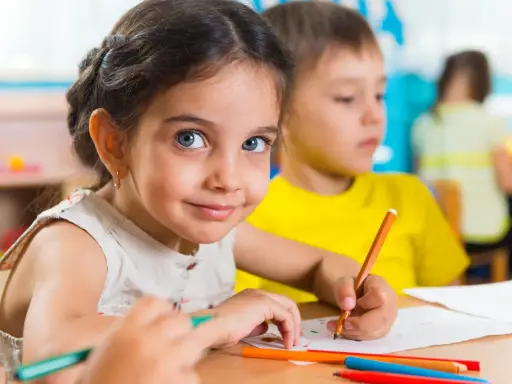 Uśmiechnięta dziewczynka z niebieskimi oczami rysuje kredką. Jej przeniesienie dziecka do innego przedszkola nie zasmuciło jej.