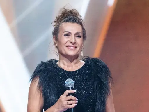Uśmiechnięta finalistka, która mogła wygrać Voice Senior, w czarnej, błyszczącej sukni z futrzanym kołnierzem, trzyma mikrofon.