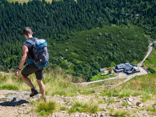 Turysta z plecakiem wspina się po kamienistym szlaku w czeskich Karkonoszach, w dole widoczny schronisko i las.