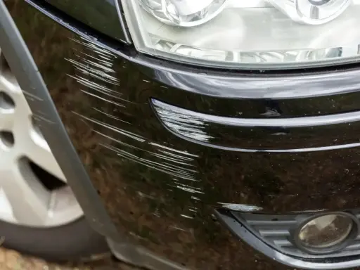 Pęknięty lakier na zderzaku czarnego samochodu, pełen licznych rys. Widać fragment reflektora i felgi.
