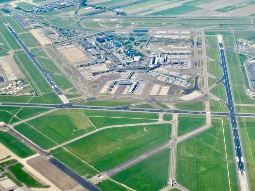Ögonblicksbild över ams flygplats med flygplan på marken och i luften, omgivet av gröna fält och vägar.