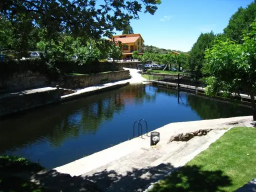 La piscina natural de Casas del Monte, rodeada de vegetación y con un edificio al fondo, invita a un refrescante baño.