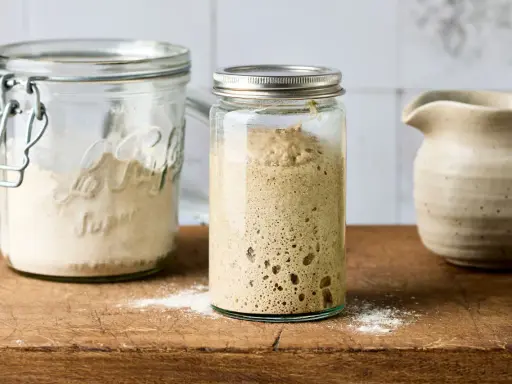 Ein Glas mit einem Sauerteigansatz, der nicht aufgeht, steht neben einem Glas mit Mehl. Ein Krug ist im Hintergrund zu sehen.