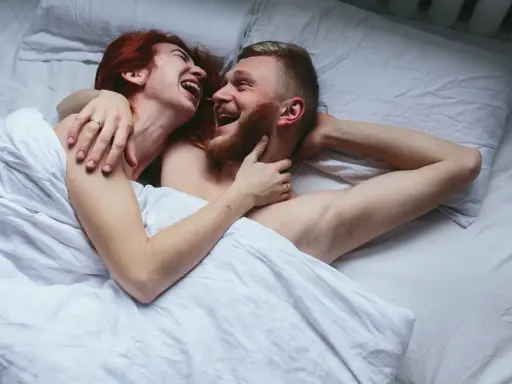 Para kochająca się w łóżku, śmiejąca się. Kobieta z rudymi włosami obejmuje mężczyznę z brodą. Sikanie podczas seksu może być częścią intymnych chwil.