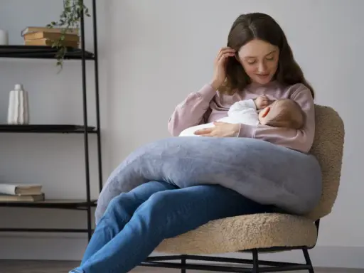 Szukasz idealny fotel do karmienia? Młoda mama z dzieckiem na rękach, otulona poduszką, siedzi na wygodnym, pluszowym fotelu.
