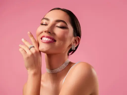 Mujer sonríe con ojos cerrados, luciendo un maquillaje romántico, joyas brillantes y un fondo rosa.