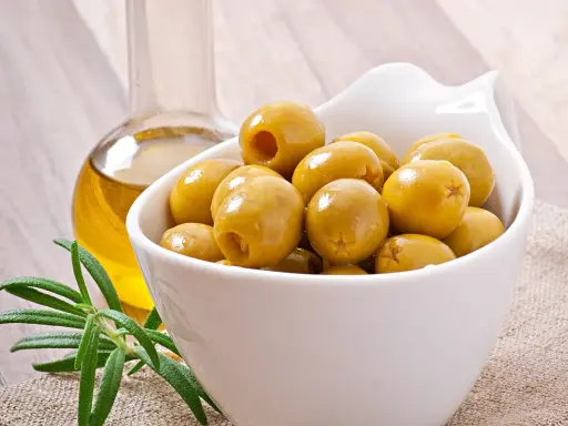 Aceitunas verdes en un cuenco blanco junto a una botella de aceite de oliva y romero. Descubre los aceitunas beneficios y contraindicaciones.