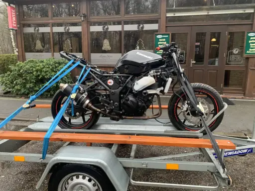 Czarny motocykl Kawasaki na przyczepce, zabezpieczony pasami. W tle budynek z napisem "Leśny Staw". Przepisy dotyczące przyczepki do motocykla są kluczowe dla bezpiecznego transportu.