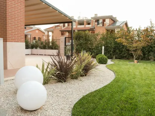 Aiuole davanti casa con ghiaia bianca, sfere luminose e prato verde curato. Sullo sfondo, case moderne e siepi.