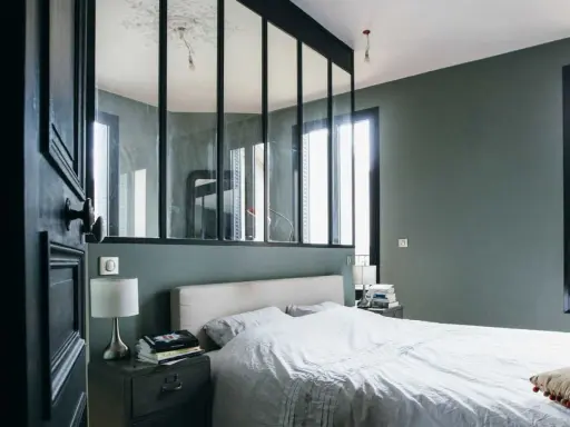 Agencement chambre moderne avec lit blanc, table de chevet industrielle et verrière séparant l'espace.