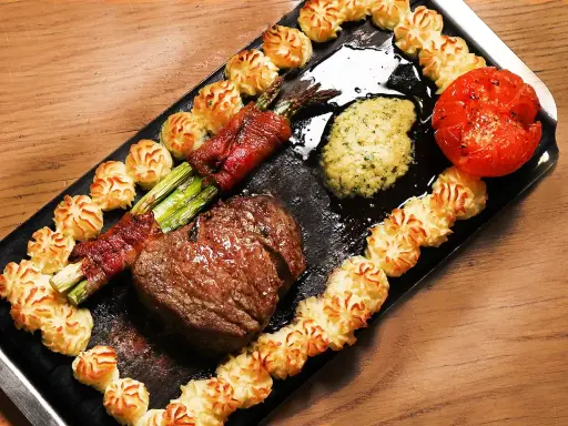 En saftig biff med sparris inslagen i bacon, serveras med potatisrosor, tomat och en grön sås. Ett perfekt plankstek recept!