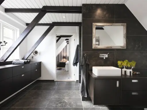 Baño moderno con vigas oscuras y suelo gris. El lavabo sobre encimera y los muebles oscuros contrastan con las paredes de baldosas oscuras. Se ve un dormitorio al fondo.