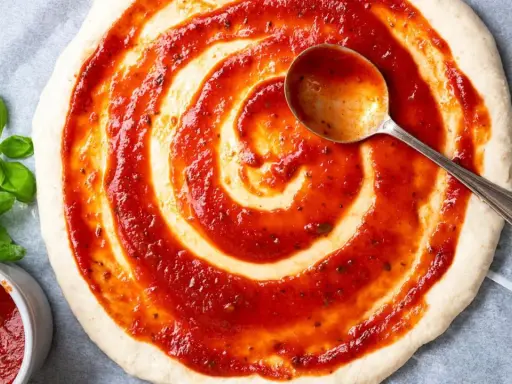 Pizzaboden mit selbstgemachter Pizzasauce bestreichen. Frisches Basilikum und ein Löffel mit Sauce daneben.