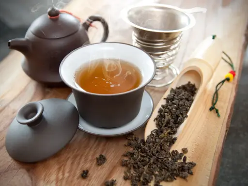 Dampfender Oolong Tee in einer Tasse, daneben Teekanne, Sieb und lose Teeblätter. Die Oolong Tee Wirkung kann belebend sein.
