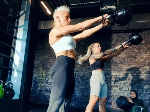 Ein Paar trainiert mit Kettlebells im Fitnessstudio. Sie heben Gewichte, während eine dritte Person im Hintergrund sitzt.
