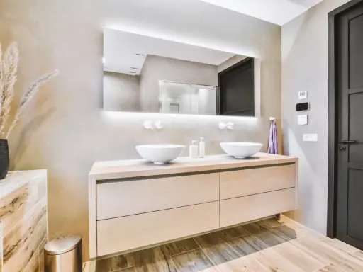 Elegantes baños modernos pequeños con lavabos dobles sobre mueble de madera clara, espejo iluminado y pared texturizada.