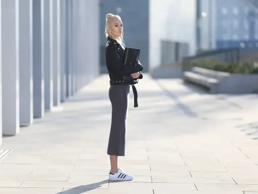 Kobieta w czarnej ramonesce i szarej spódnicy prezentuje stylowe adidas superstar stylizacje na tle nowoczesnej architektury.