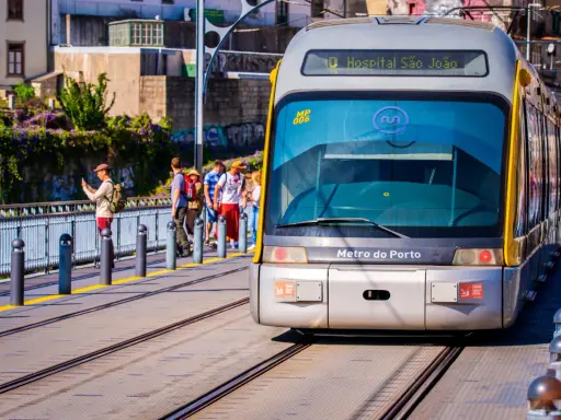 En modern spårvagn från Metro do Porto närmar sig stationen. Passagerare väntar på perrongen, omgivna av grönskande växtlighet.