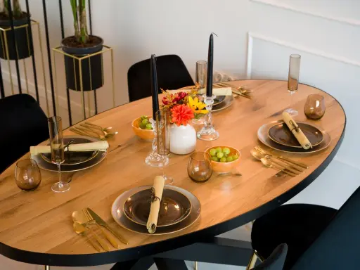 Un dîner élégant est prêt, avec des assiettes sombres, des couverts dorés et des raisins verts. Les art de la table sont mis en valeur par des bougies noires et des fleurs colorées.