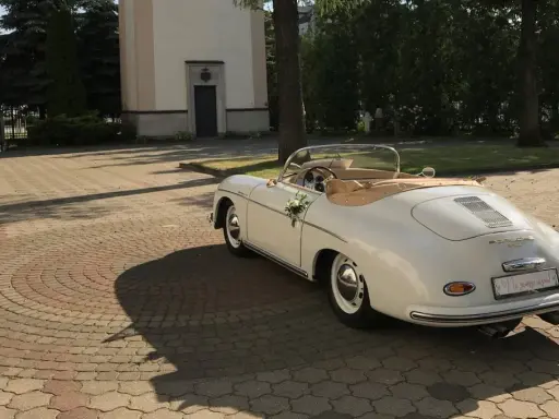 Biały, zabytkowy kabriolet Porsche, udekorowany kwiatami, czeka przed kościołem. To idealny samochod na wesele, gotowy na podróż poślubną.