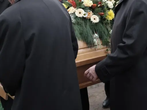 Mężczyźni w czarnych płaszczach niosą trumnę ozdobioną kwiatami. Kondukt pogrzebowy w żałobnej ciszy.