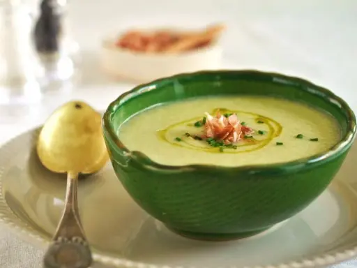 Un refrescante gazpacho de melón, adornado con jamón serrano y cebollino, servido en un cuenco verde con una cuchara dorada.