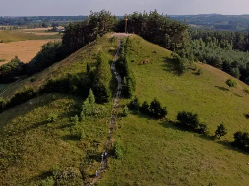 Wzgórze porośnięte trawą i drzewami, ze schodami prowadzącymi na szczyt, gdzie stoi drewniany krzyż. Widok na pola i lasy.