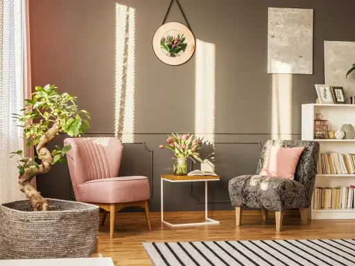 Un angolo relax con due poltrone, un tavolino con fiori e libri, una pianta e una libreria. La luce del sole crea un'atmosfera accogliente.