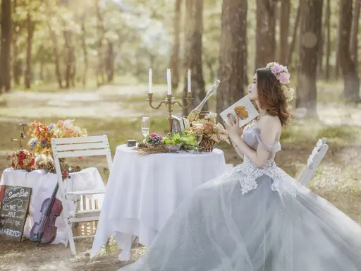 Romantyczna ceremonia ślubna w lesie. Panna młoda w pięknej sukni czyta książkę przy stole zastawionym owocami i szampanem.