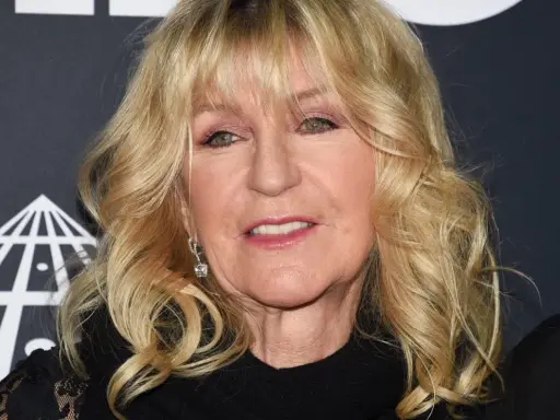 Christine McVie, en ikonisk sångerska i Fleetwood Mac, ler med sitt blonda hår och smycken.