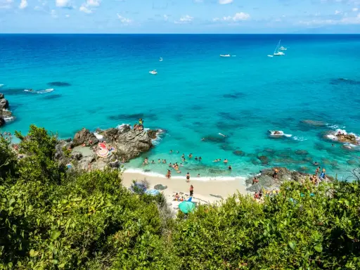 Spiaggia affollata con gente che nuota e prende il sole, barche in mare e vegetazione lussureggiante. Un angolo di paradiso del **calabria mare**.