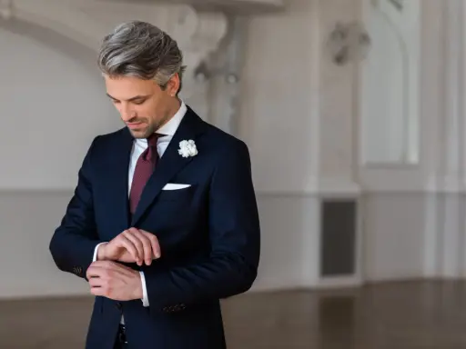 Elegancki mężczyzna w granatowym garniturze i białej koszuli, z bordowym krawatem i butonierką, przygotowuje się na wesele.