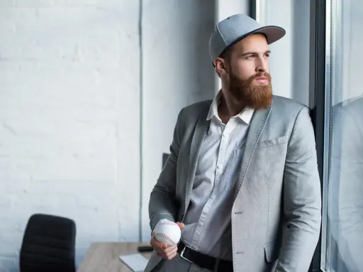 Mężczyzna w szarej czapce snapback i garniturze trzyma piłkę baseballową.