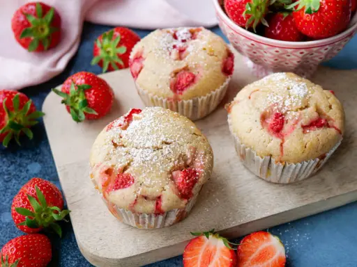 Saftige Erdbeer Muffins, bestäubt mit Puderzucker, liegen auf einem Holzbrett, umgeben von frischen Erdbeeren.