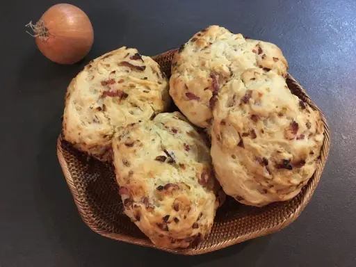 Quatre pains aux lardons et oignons dorés dans un panier en osier, avec un oignon cru à côté.