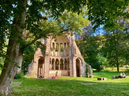 Ruinen einer alten Kirche in NRW, umgeben von Bäumen. Menschen genießen ihre Freizeit auf der grünen Wiese.