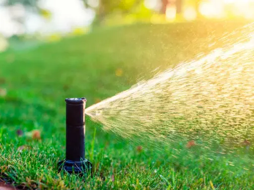 Aspersor de riego automático en acción, rociando agua sobre césped verde bajo la luz del sol.