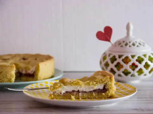Slice of crostata with ricotta and jam on a yellow polka dot plate, with a whole crostata in the background.
