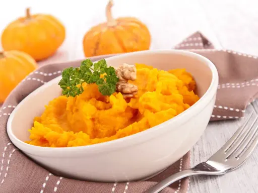 Un tazón de cremoso **puré de calabaza al horno**, adornado con perejil y nueces. Al fondo, calabazas pequeñas y una servilleta marrón.
