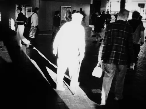 A stark black and white image by Trent Parke captures a blurred figure in a white hat and coat, walking with a cane. Other figures are also blurred, suggesting movement and a fleeting moment.