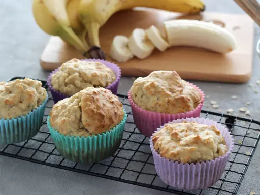 Fünf leckere bananenmuffins ohne zucker in bunten Förmchen auf einem Gitter. Bananen und Mehl im Hintergrund.