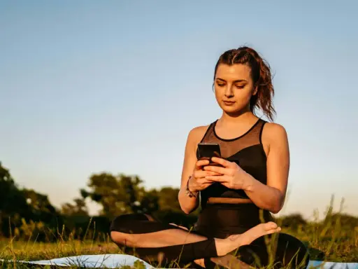 Kvinna i lotusställning på en matta utomhus, med en mobiltelefon i händerna. En modern typ av meditation.