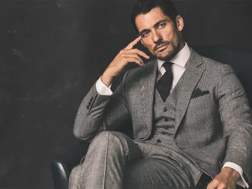 A man in a grey suit, black shirt, and tie, with a thoughtful expression, sits in a chair.
