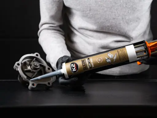 Ręce w czarnych rękawiczkach aplikują czarny silikon K2 na część pompy wodnej. Zastanawiasz się, ile schnie silikon? Ten produkt jest odporny na wysokie temperatury.