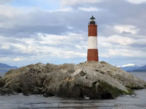 Faro a strisce rosse e bianche su un'isola rocciosa nel Canale di Beagle, con montagne innevate sullo sfondo.